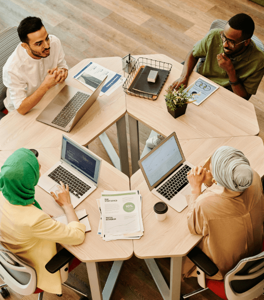 A diverse team of professionals collaborating at a round table with laptops and documents in a modern office setting.